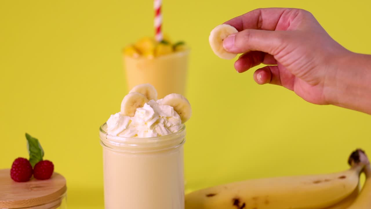 Hand places banana slices on creamy milkshake with whipped cream, bright yellow background, studio lighting