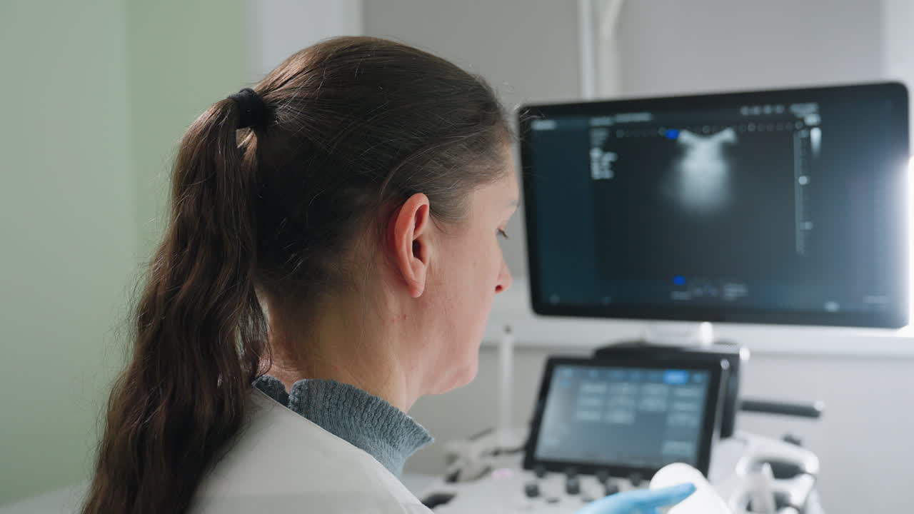 Medical laboratory scientist applies gel on transducer while maintaining strong focus on monitor displaying internal scan results in clinical environment, showing precision during diagnostic process