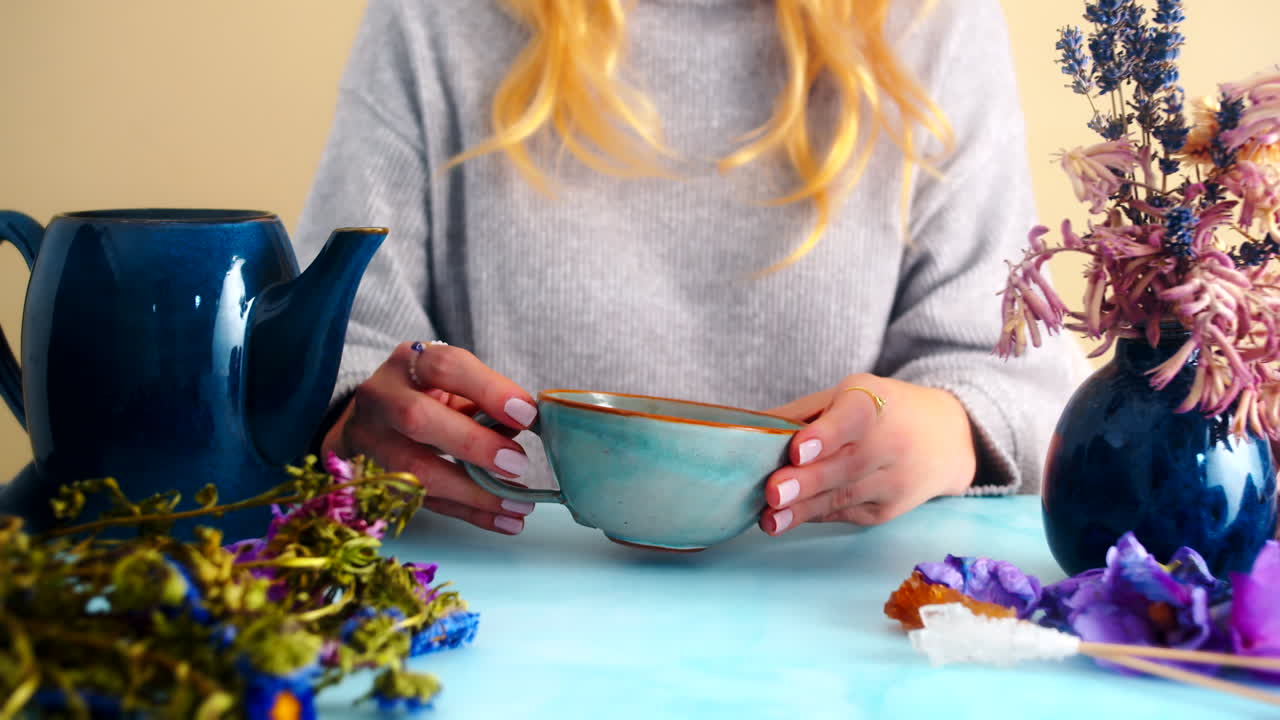 Female hands holding and drinking from a ceramic tea cup in a cozy setting with a blue teapot and dried flowers. Ideal for lifestyle, tea rituals, or relaxing moments with a calm, natural vibe.