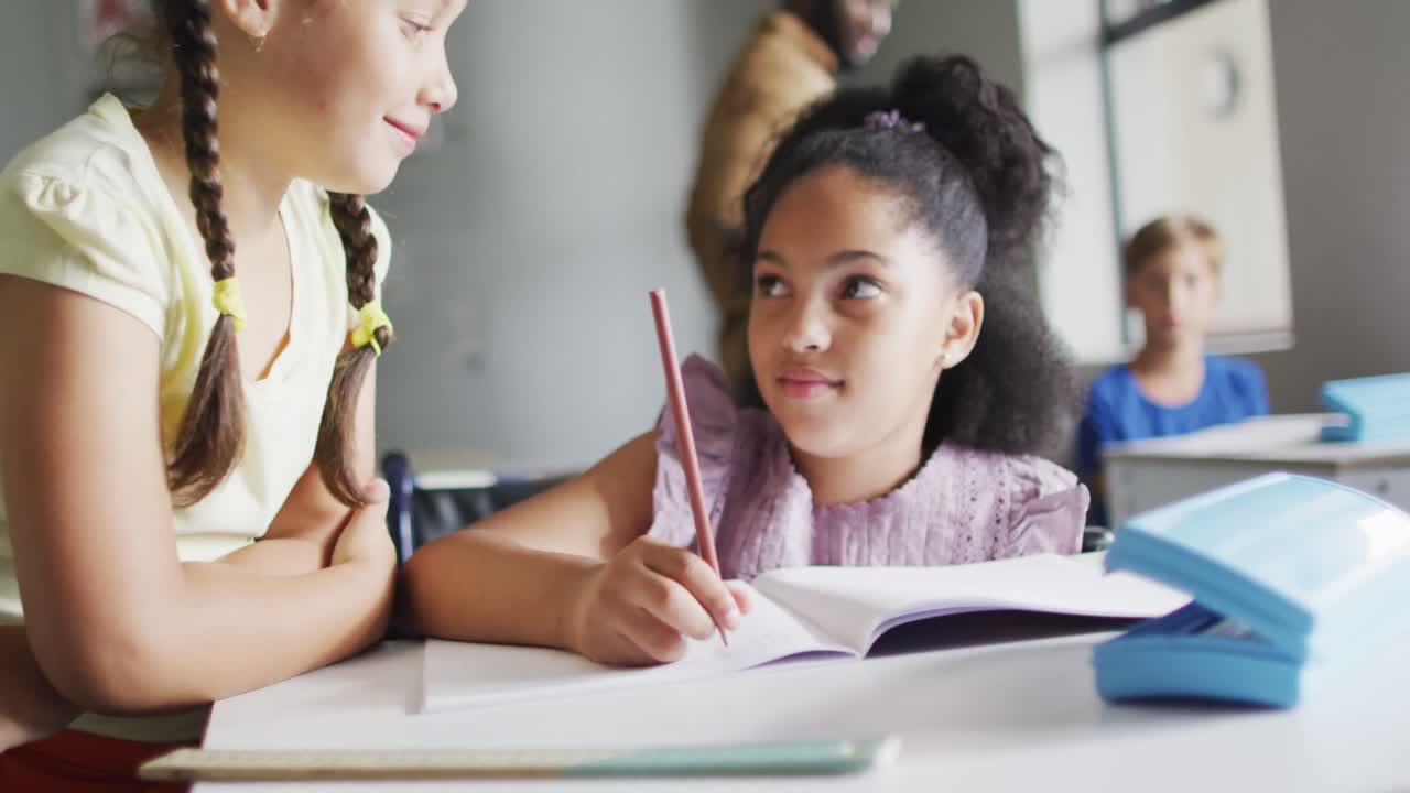 Video of happy diverse girls at desk doing lessons together in classroom