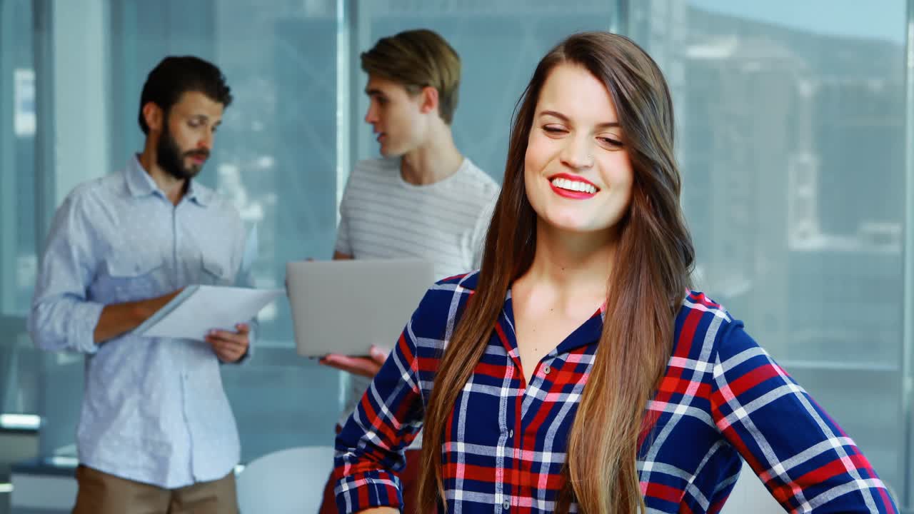 Executiva sorridente de pé com as mãos na cintura enquanto colegas discutem ao fundo.
