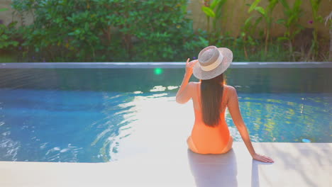 Vista-Trasera-De-Una-Mujer-Sentada-En-El-Borde-De-La-Piscina-Con-Traje-De-Baño-Naranja-Y-Sombrero