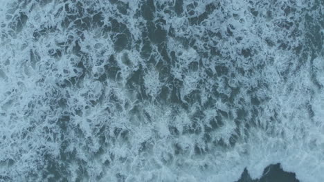 an overhead drone shot captures the textures of water as sea waves crush, foam, bubble, and swirl