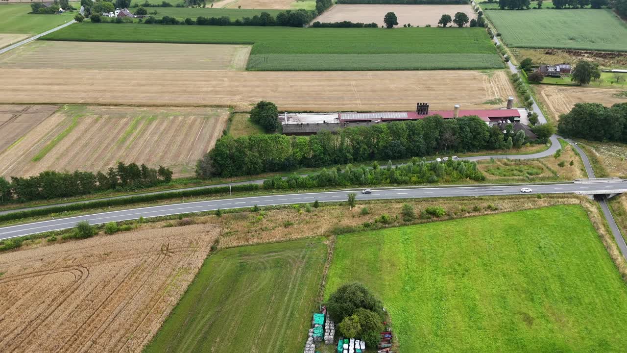 Aerial view of rural farmland with harvested fields, a country road and farm building surrounded by green landscape under a cloudy sky. Lateral shot. Traffic with cars on street.Summer day on America
