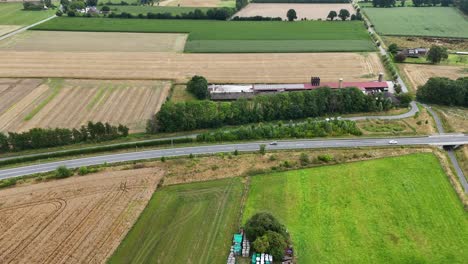 aerial view of rural farmland with harvested fields, a country road and farm building surrounded by green landscape under a cloudy sky. lateral shot. traffic with cars on street.summer day on america
