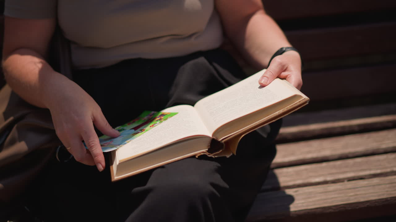 Clos up woman sitting on wooden bench outdoors turning new page of old book under warm sunlight, focusing on hands and soft shadows, peaceful atmosphere of leisure reading in summer afternoon