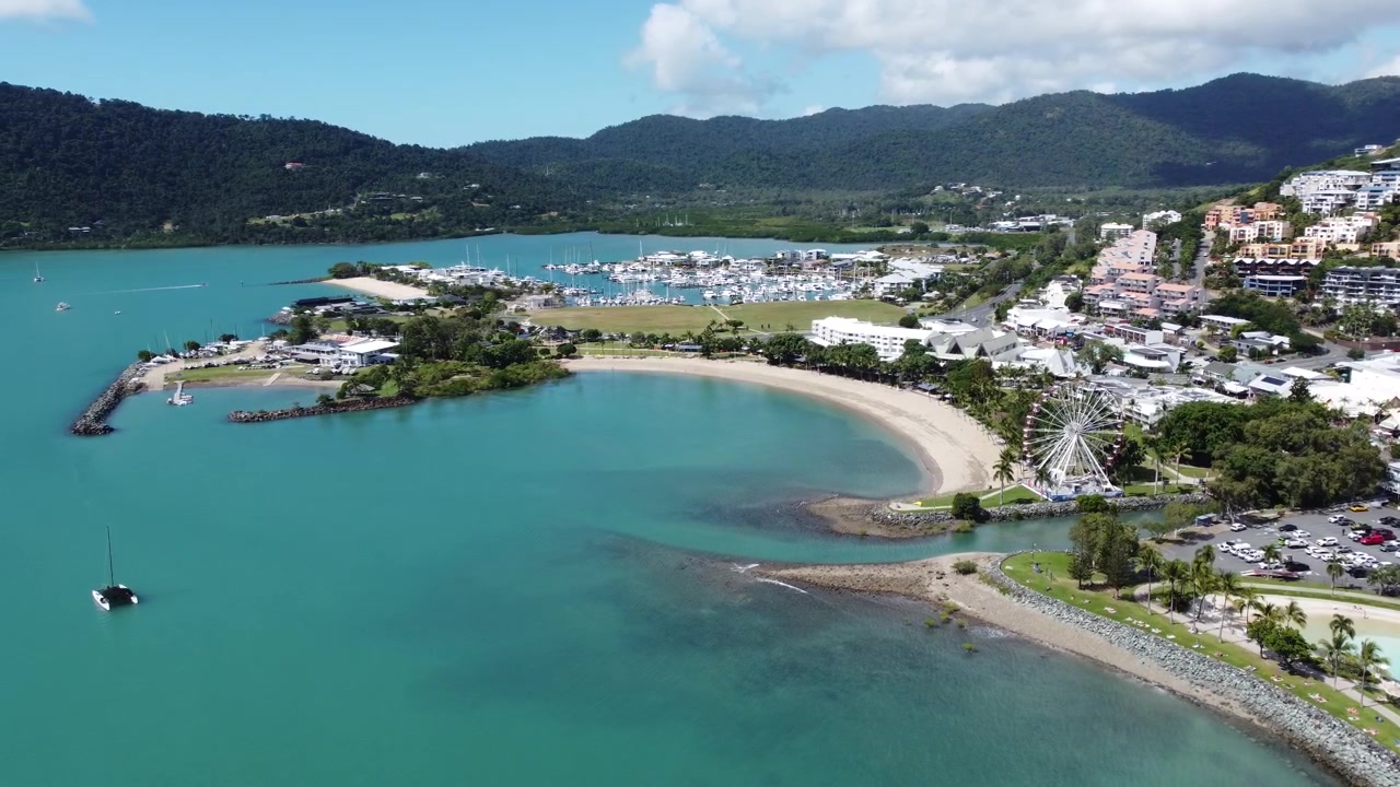 Prospettiva aerea dall'alto del litorale di Airlie Beach che cattura la ruota panoramica, la baia sabbiosa, il porto turistico e lo sfondo montuoso in una giornata limpida