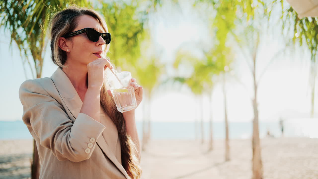 Elegant woman holding a glass of lemon water while relaxing at a beach bar under palm trees