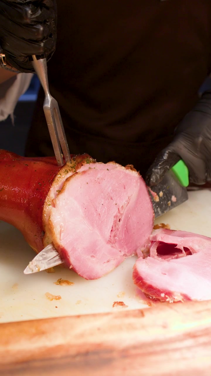 Primo piano di uno chef con guanti neri che intaglia prosciutto arrosto con coltello e forchetta.
