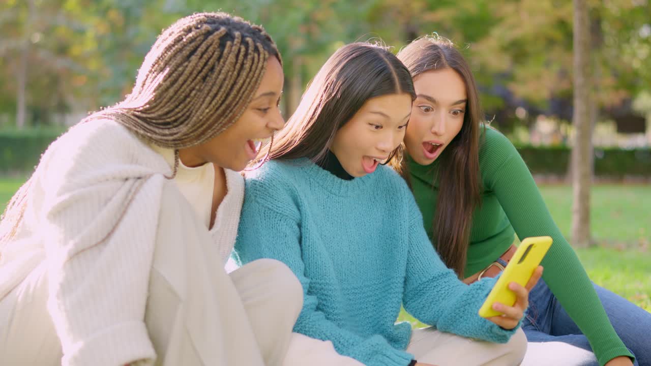 Three diverse young women react with surprise while looking at a smartphone in a park