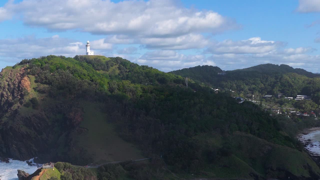 Aerial footage of Byron Bay lighthouse atop a lush hill, under a partly cloudy sky, with coastal views