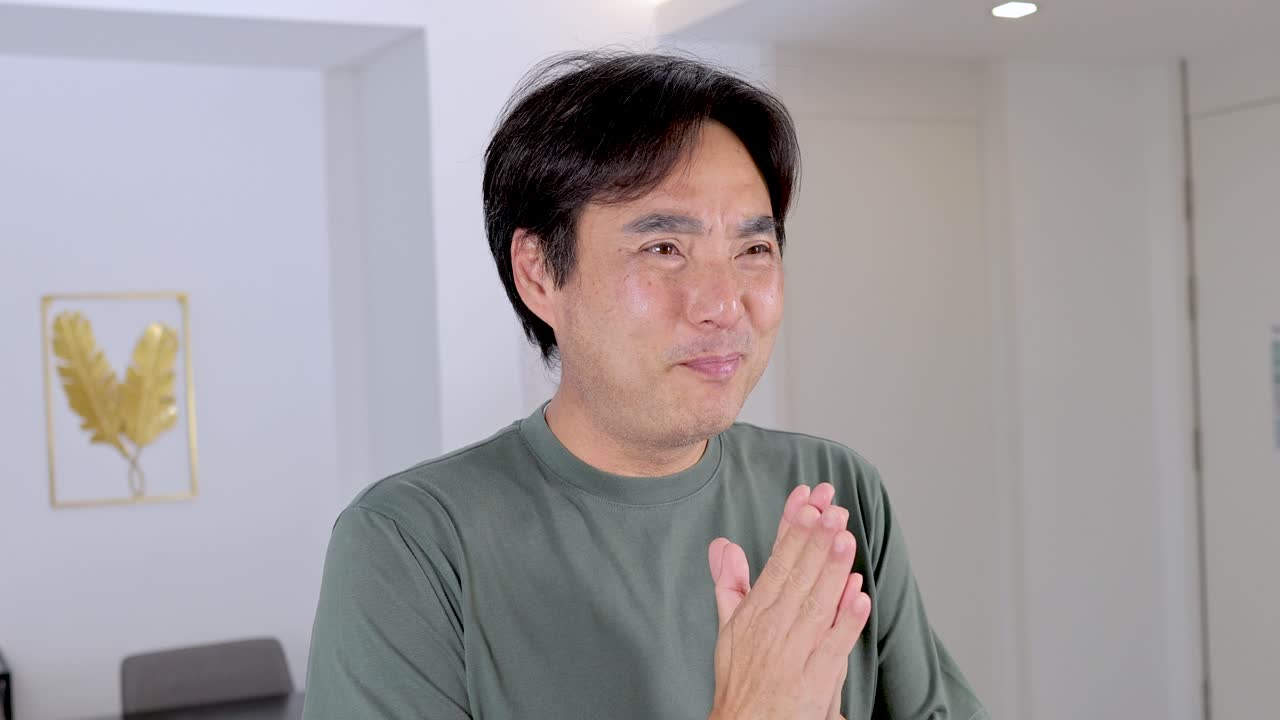 Asian man smiles warmly, hands together in polite greeting gesture, bright indoor setting, steady camera
