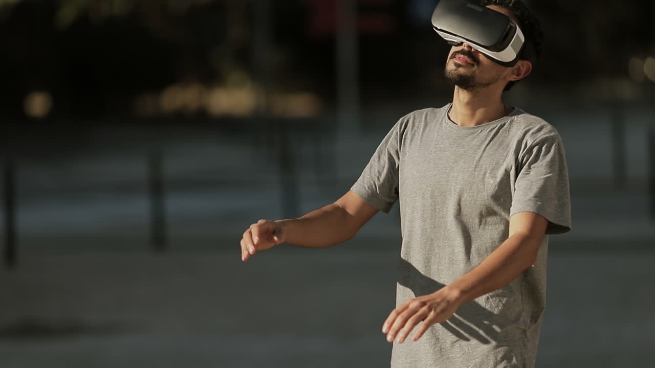 joven árabe con cabello rizado oscuro y barba en camiseta gris estando en el parque con gafas de realidad virtual