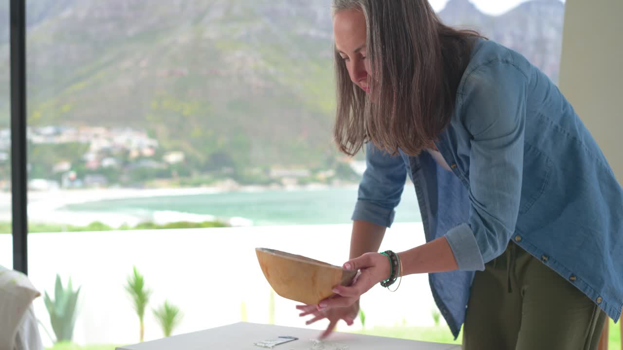 Mature adult woman inspecting underside, tilting gourd at small white table by bay window to drain
