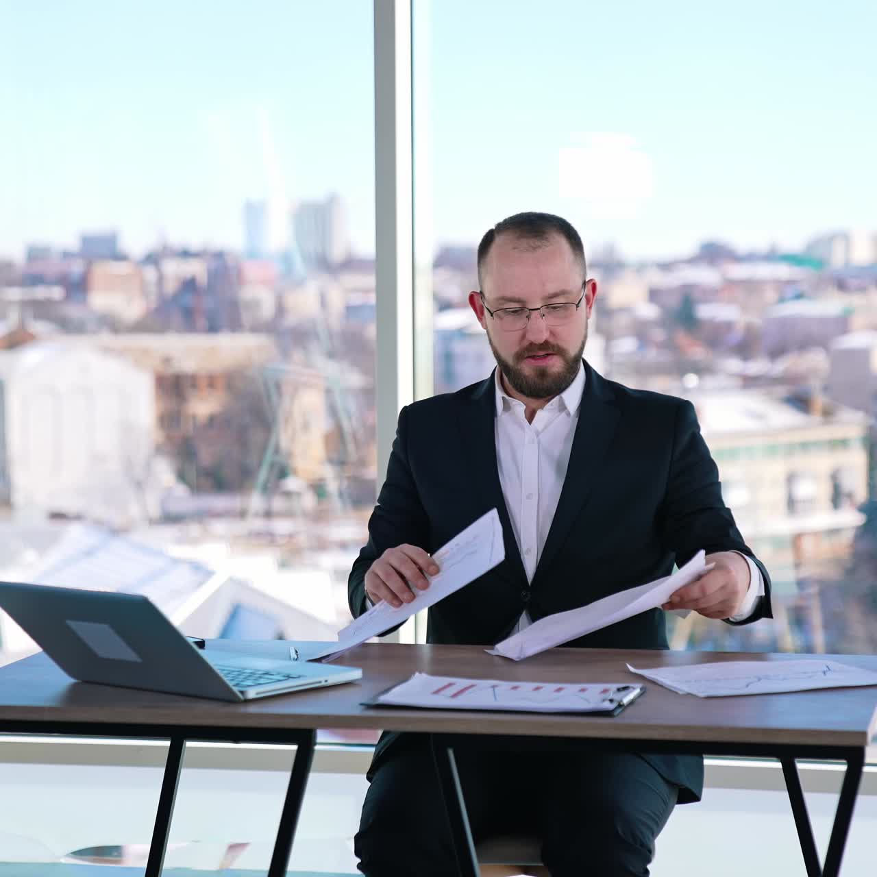 Portrait of a businessman at workplace. Bearded office worker in suit sitting at the table with a laptop on window background in the city
