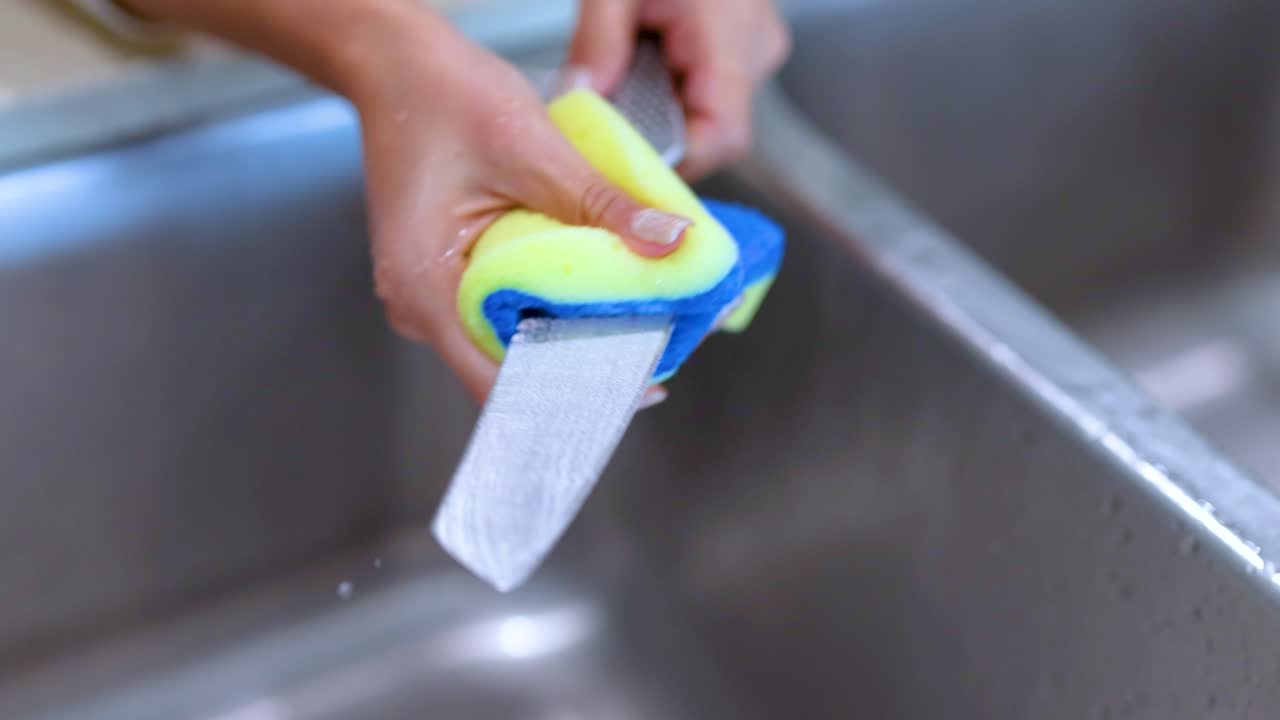 A person cleans a knife with a sponge in a stainless steel sink, emphasizing hygiene and kitchen maintenance