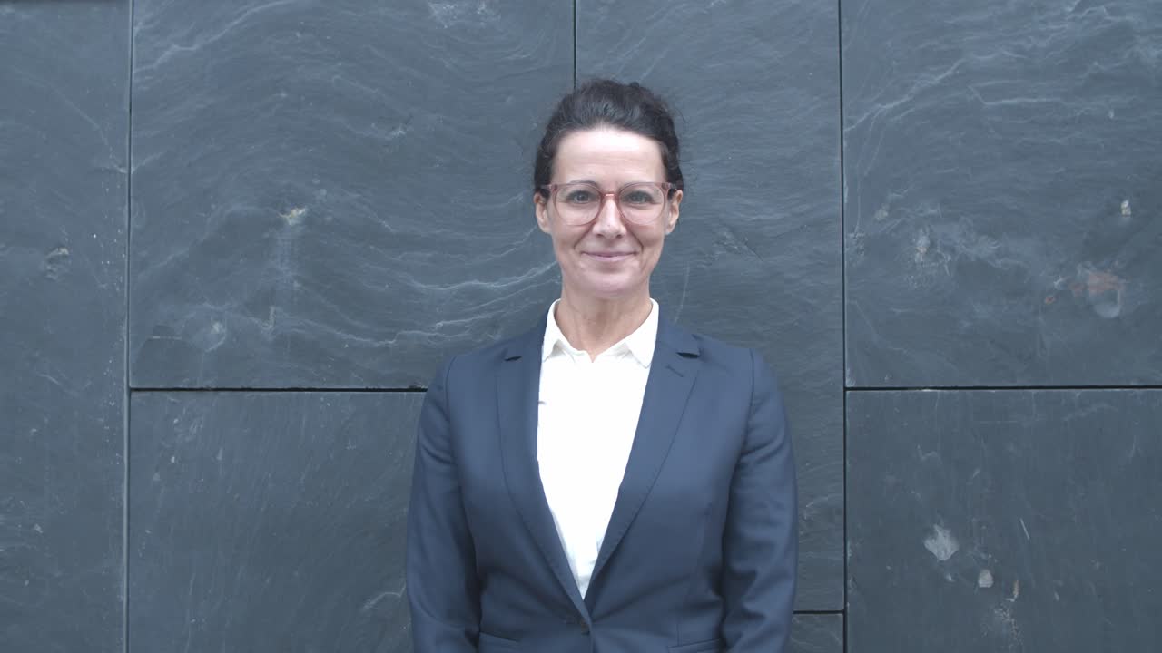 Positive middle aged Caucasian businesswoman in suit and glasses standing at outdoor building wall, posing and smiling at camera