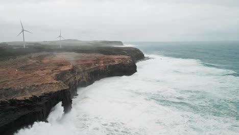 Vista-Aérea-Delantera-De-Las-Mareas-Oceánicas-Golpeando-Las-Rocas-De-La-Costa-De-Cape-Bridgewater,-Australia,-Con-Un-Parque-Eólico-Al-Fondo