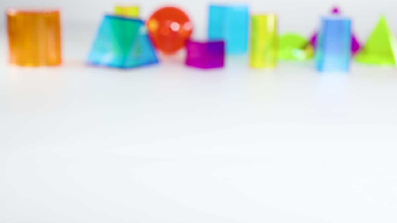 A hand places a red plastic pyramid among colorful geometric shapes on a white table. Bright lighting highlights the scene