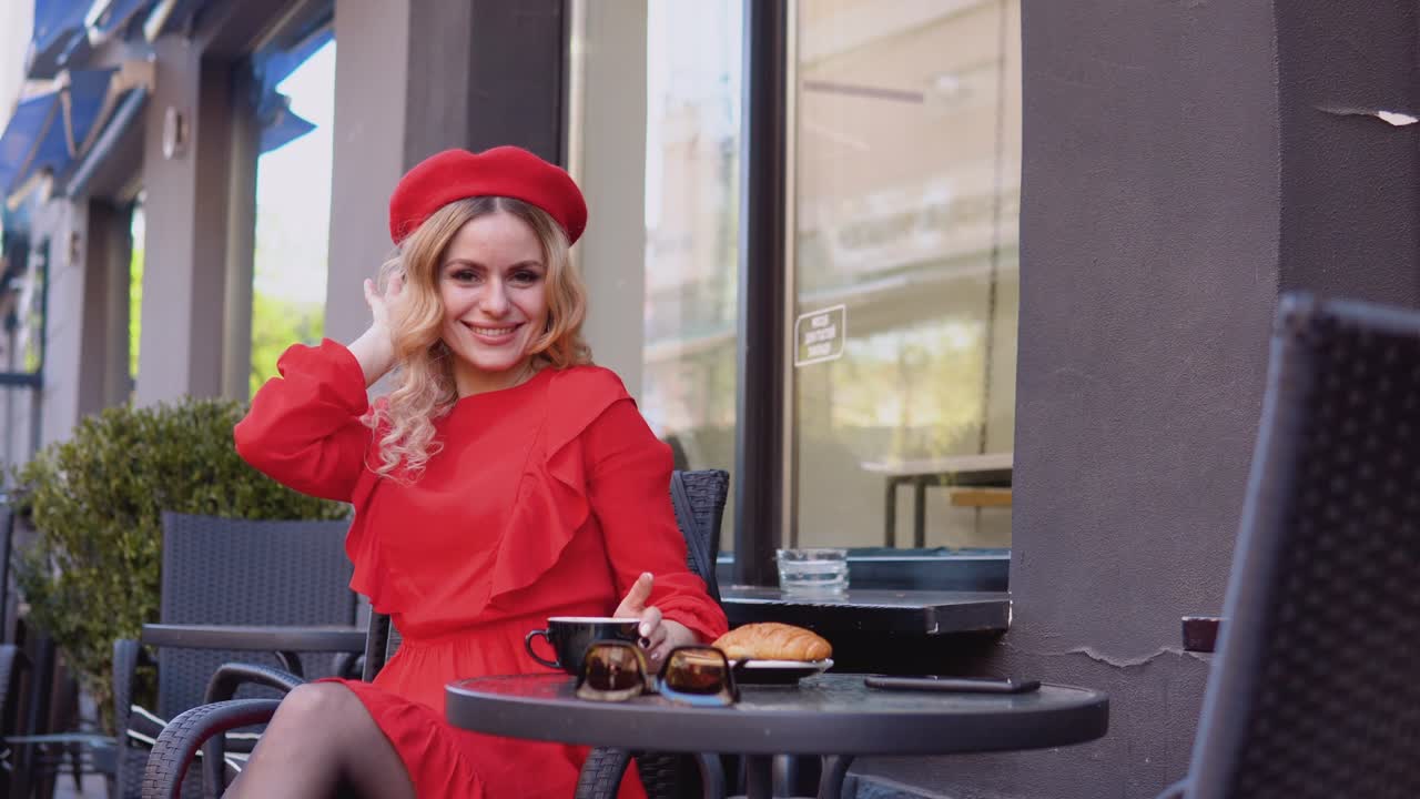 Young romantic attractive woman smiling looking at camera. Woman in a red dress and beret sitting with a cup of coffee at a table on the street