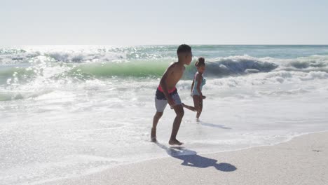 Lächelnde-Afroamerikanische-Geschwister-Laufen-Und-Tragen-Badeanzüge-Am-Sonnigen-Strand