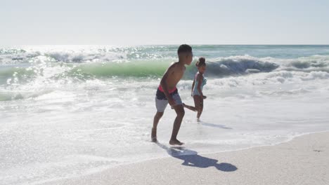 hermanos afroamericanos sonrientes corriendo y usando trajes de baño en una playa soleada