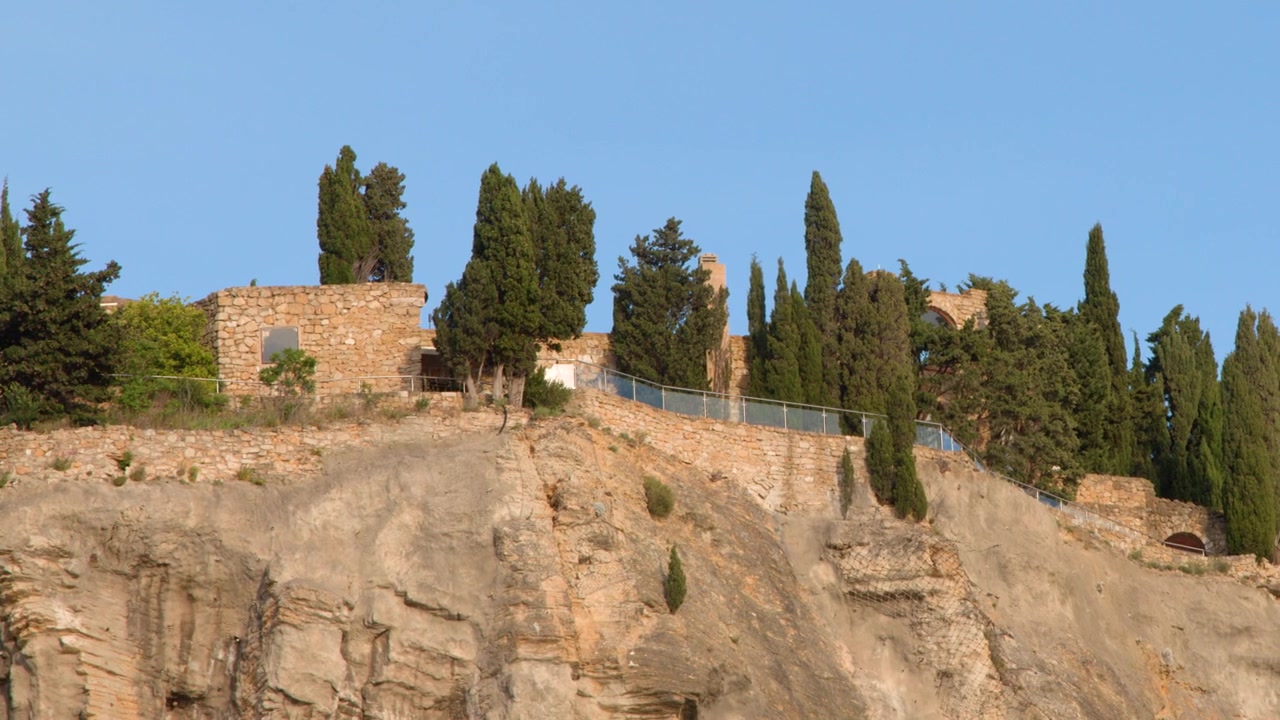 Inquadratura ampia statica di un castello medievale in pietra e cipressi su una scogliera rocciosa.