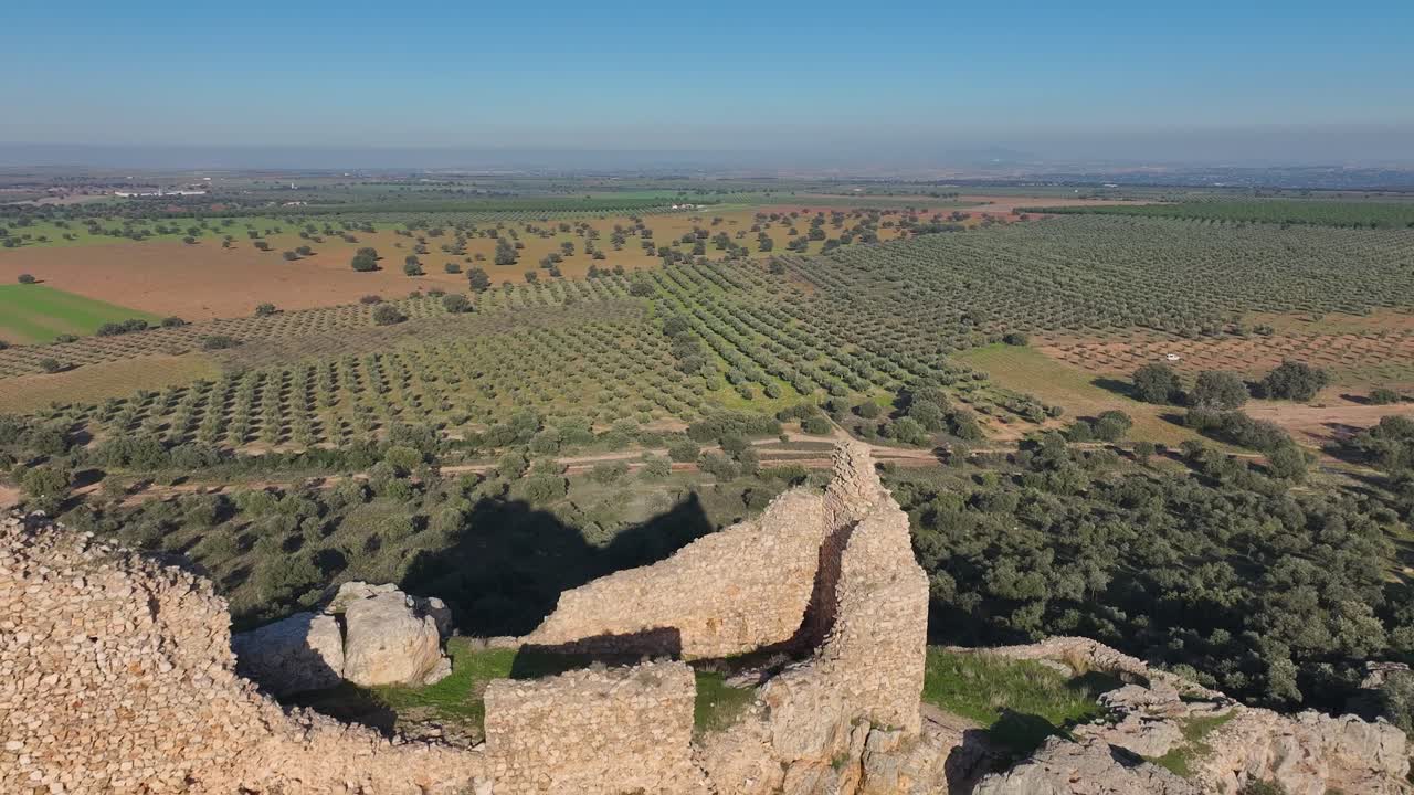 Aerial drone shot capturing the full ruins of Dos Hermanas Castle on a rocky hill in Navahermosa, Toledo. Surrounded by olive groves, farmland, and distant mountains under a clear sky