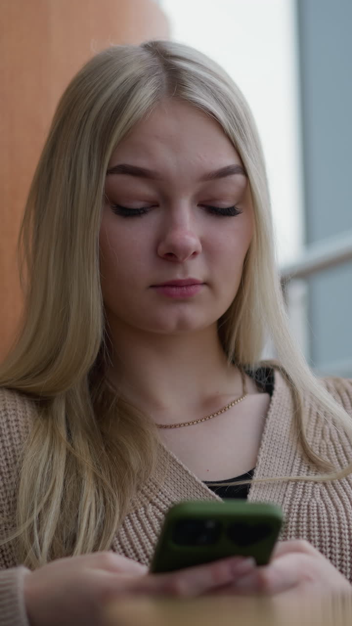Jovem mulher com um colar no pescoço, sentada e segurando um telefone, com um olhar pensativo e uma vista desfocada da cidade visível através de uma janela, em uma atmosfera moderna e relaxada em um ambiente urbano.