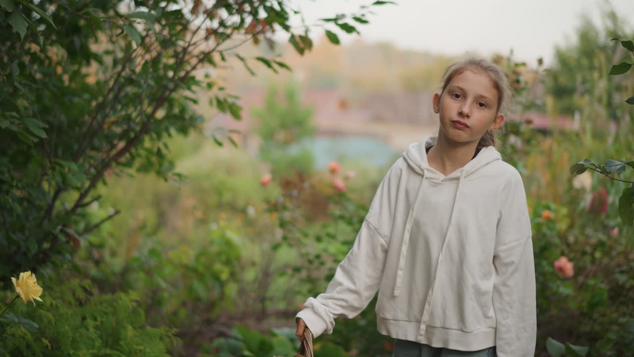 Nahaufnahme einer jungen Frau im Garten, sanfter, nachdenklicher Ausdruck, Hoodie-Detail, geflochtenes Haar umrahmt das Gesicht, Hände auf einem Korb, nachdenkliche Stimmung, Naturstudentin, Träumerin, Dichterin-Persona, geringe Tiefenschärfe, warmes Abendlicht.