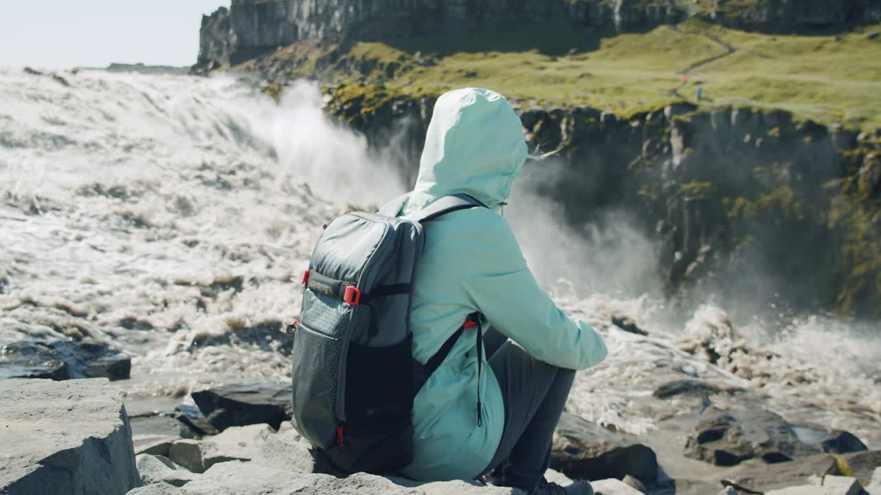 mujer sentada en el borde de un acantilado. río en flujo desfocalizado cayendo en la cascada de detifoss en el fondo. iceland