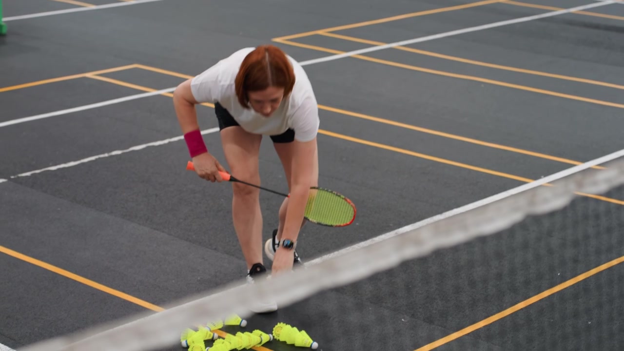 Hvit kvinne praktiserer badminton nær nettet, samler fjærballer på innendørsbane, fritidsspiller som forbedrer kortspillet, iført hvit t-skjorte og rosa armbånd, jevnt tempo, fokusert uttrykk.