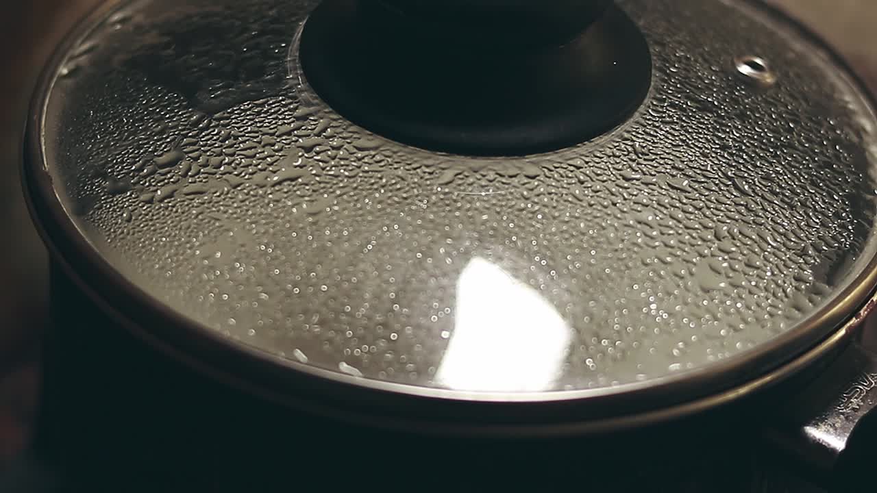 A close-up view of a black cooking pot with a glass lid covered in condensation from steam showing daily chores and peaceful home life