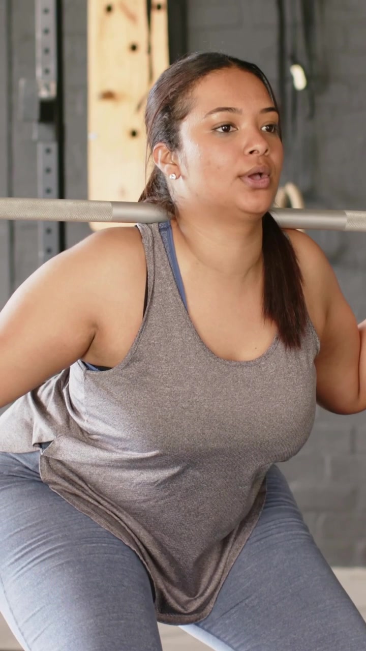 Video verticale: Donna che espira mentre fa uno squat con bilanciere in palestra, con bilanciere e rack, in canotta grigia per la forza.