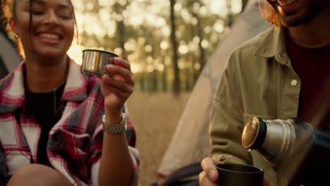 Close-up-shot-of-a-brunette-man-in-a-light-green-jacket-pouring-tea-from-a-thermos-of-a-girl-with-Black-skin-in-a-checkered-shirt-during-their-hike-in-a-light-green-sunny-summer-forest