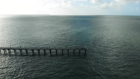 Ariel-pullback-of-the-end-of-a-wooden-pier-that-stretches-into-the-vast,-dark,-gloomy,-blue-ocean-on-a-sunny,-afternoon-day-where-the-fluffy,-white-clouds-float-overhead-in-the-gorgeous-blue-sky