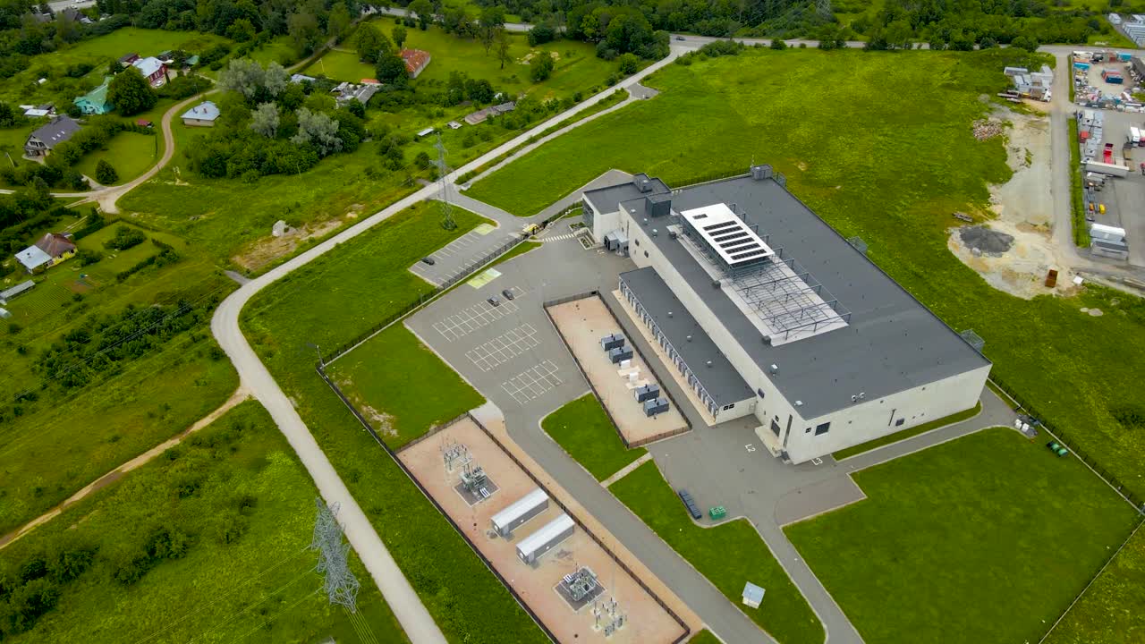 Aerial drone footage orbiting around a gray colored server room or data centre in green grassy countryside that is fenced and has roads around it. Cooling system on the rooftop to cool the server room