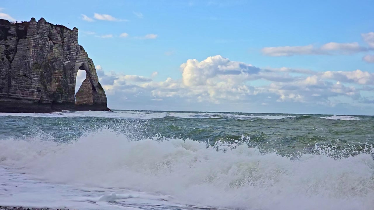 Slow motion waves reaching the shore with Etretat cliffs and natural arch in Normandy France under blue sky