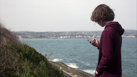 joven adolescente buscando en las redes sociales en su teléfono móvil con vista costera en segundo plano, copiando espacio