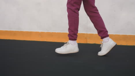 female coach jogging on curved asphalt court beside barrier wall with yellow edge, close up of legs clad in purple tracksuit and white sneakers stepping on sports surface under soft pastel dusk light