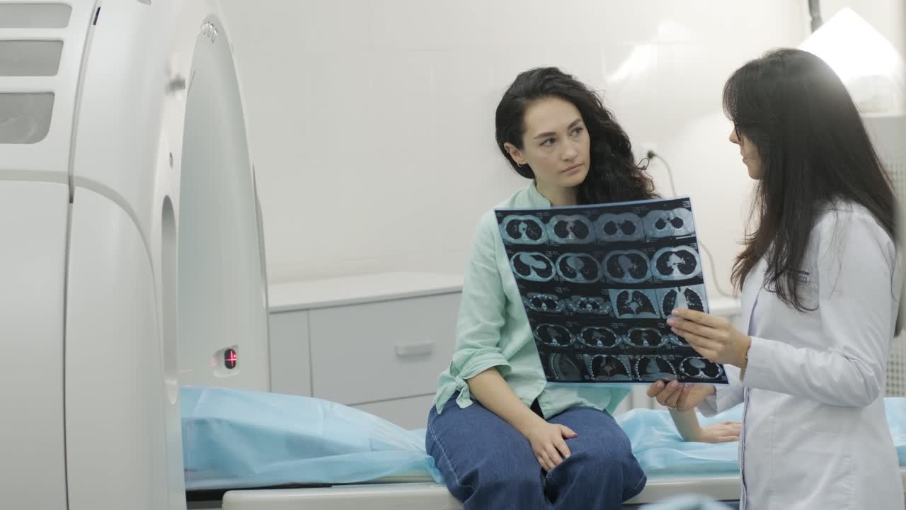 Woman doctor radiologist explains the results of CT scanning for young female patient, showing the snapshot with images, observing and analyzing CT scan in modern clinic beside modern CT scanner