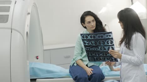 woman doctor radiologist explains the results of ct scanning for young female patient, showing the snapshot with images, observing and analyzing ct scan in modern clinic beside modern ct scanner