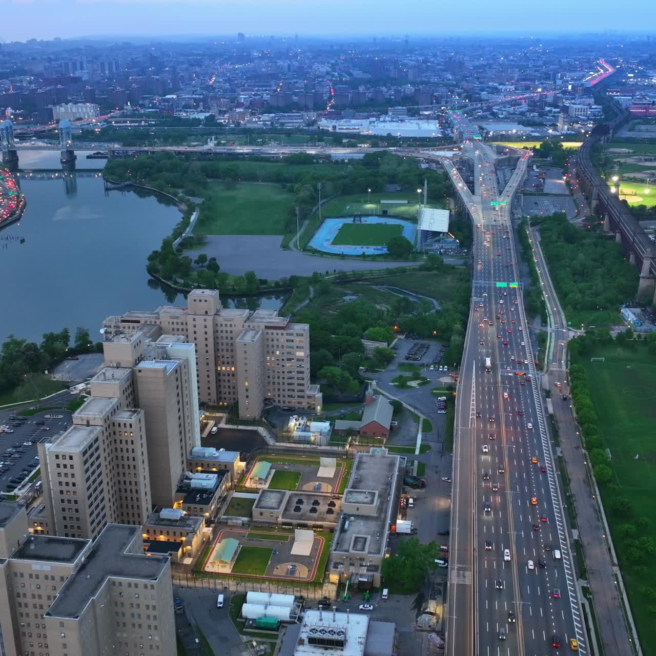 Speed motorway with lots of transport passing by. Drone footage over the city with green parks, rivers and stadiums. New York at sun down