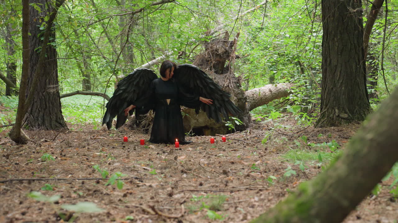 Magician dressed in flowing dark gown stands before five glowing candles on forest ground, pausing in stillness before kneeling to perform sacred prayer to ancient gods