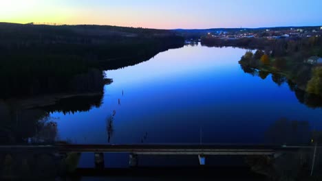 Imágenes-De-Drones-Que-Revelan-Un-Puente-De-Tren-En-Una-Hermosa-Tarde-Con-Un-Lago-Tranquilo-En-Primer-Plano-Y-La-Puesta-De-Sol-En-El-Fondo