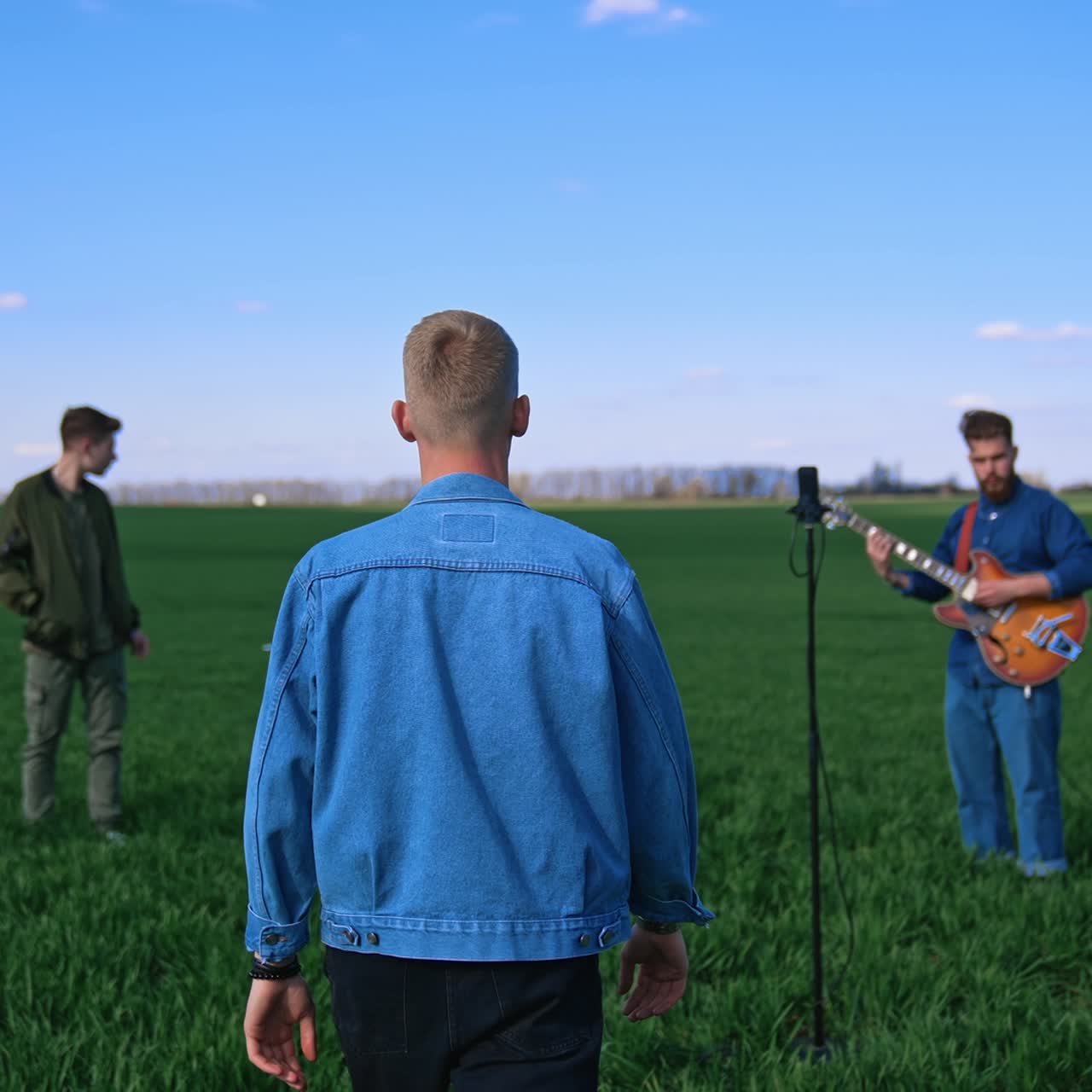 Music band on green field background. Young men playing musical instruments outdoors. Man singer coming to a microphone
