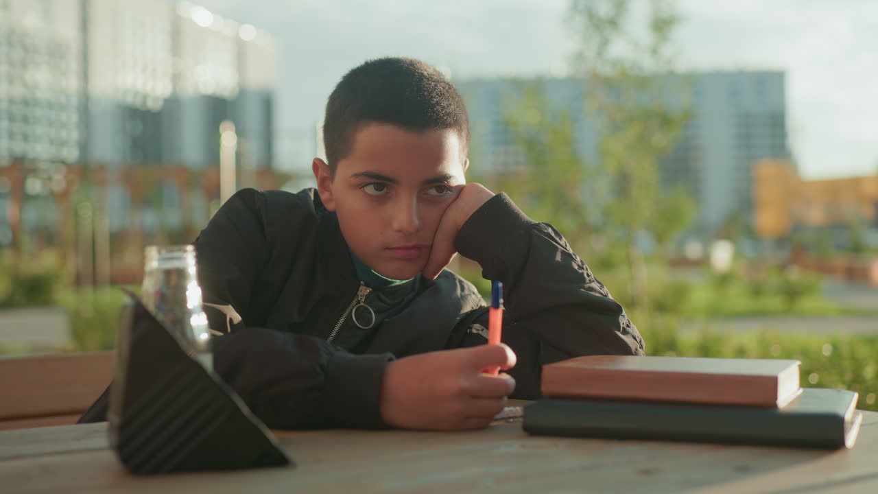 Boy lost in thought writing on wooden table with notebook and pen while hand offers snack and drink, blending study concentration with comfort in supportive outdoor environment