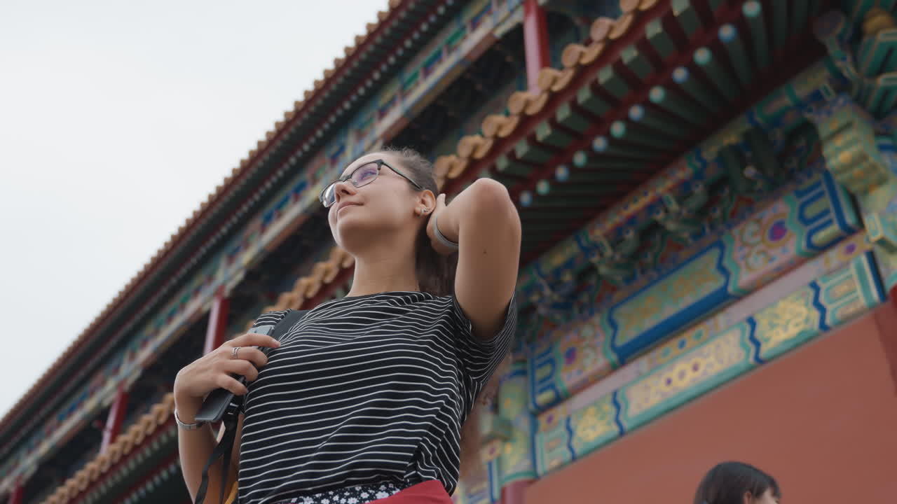 Mujer de Asia Oriental bajo un ornamentado templo chino, con aleros del techo y ménsulas pintadas, mirando hacia arriba con gafas, mano en la cadera, camisa de lunares y falda roja, ambiente de turista urbano, cielo nublado, toma de ángulo bajo.