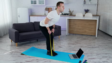 concentrated man exercising with a rubber band at home. young man doing working out while standing on a mat in the kitchen. sportive lifestyle during quarantine