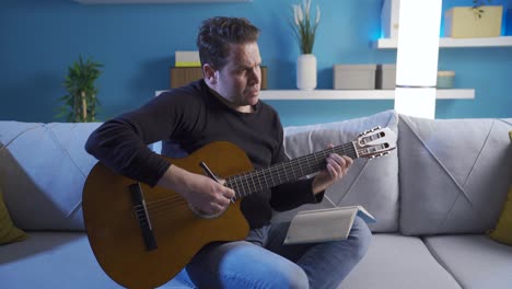 male musician sitting on sofa at home, playing guitar and writing lyrics in notebook while composing new song.